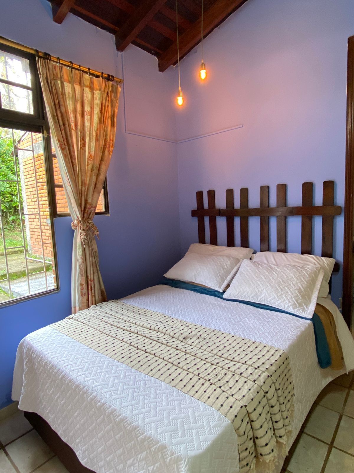 A bedroom with a bed, curtains, a wooden headboard, and a window with a view of greenery.