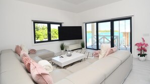 A modern living room with a large sofa, a coffee table, and a flat-screen TV. There are large windows with a view of the sea.