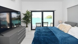 A modern bedroom with a large bed, a TV, and a balcony with a view of the sea.