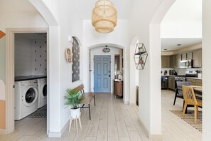 A laundry room with a washing machine, a bench with a potted plant, a blue door, and a kitchen area with cabinets and a dining table. - Flower Street House: Pool, Spa, Golf! (Queen Creek)