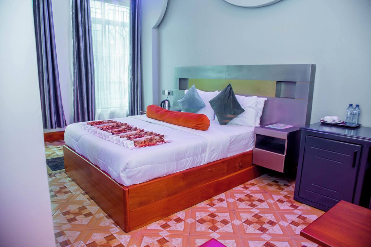 Quarto de hotel com uma cama grande, mesa de cabeceira, guarda-roupa e vista para o exterior pela janela.