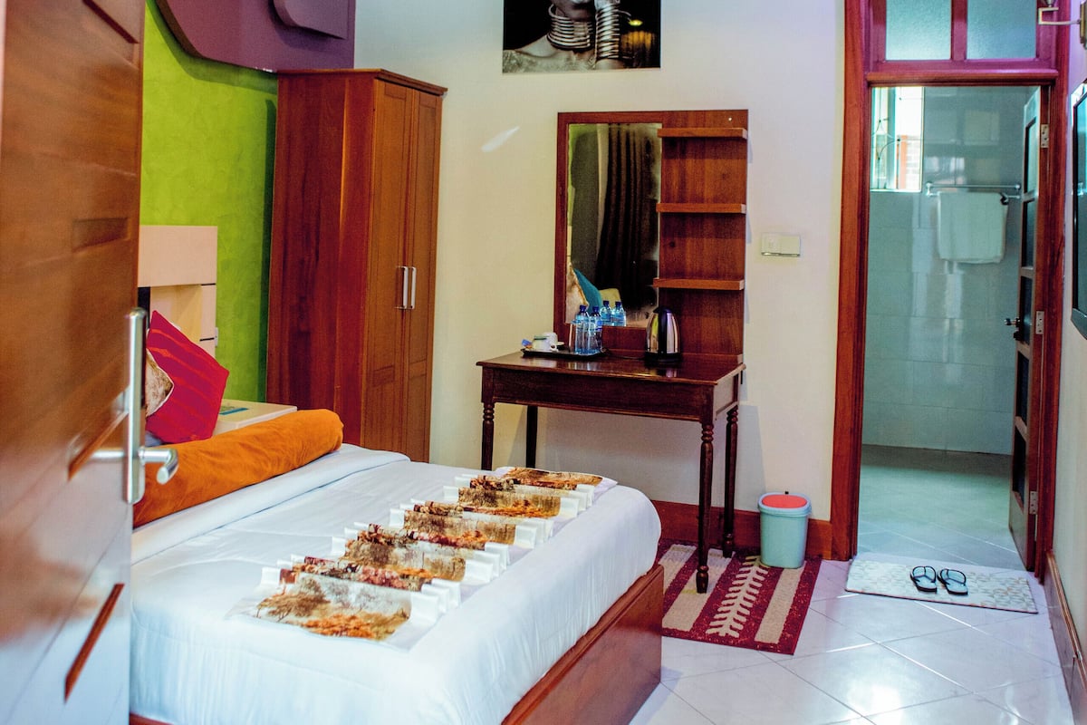 Quarto com cama de madeira, uma escrivaninha com cadeira, espelho e porta para um banheiro.