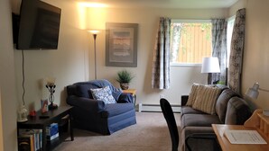 Ein Wohnzimmer mit einer Couch, einem Couchtisch, einem Fernseher, einer Lampe und einem Fenster mit Vorhängen.