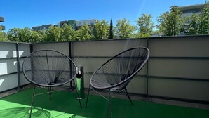 Dos sillas de estructura metálica negra sobre un área de césped artificial verde, con un muro metálico y edificios al fondo.