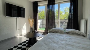 Un dormitorio con una cama grande, un televisor de pantalla plana en la pared, una mesita redonda y vistas al exterior a través de una ventana con cortinas traslúcidas.