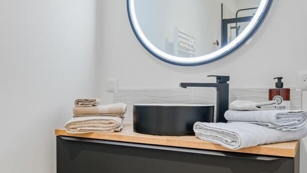 Un baño con un espejo redondo, un lavabo negro y un tocador de madera con toallas dobladas.