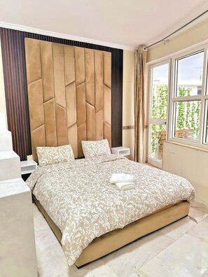 A bedroom with a large bed, a headboard, bedside tables, and a window with a view.