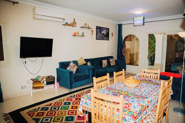 A living room with a blue sofa, a TV, a patterned rug, a dining table with chairs, and a small side table.