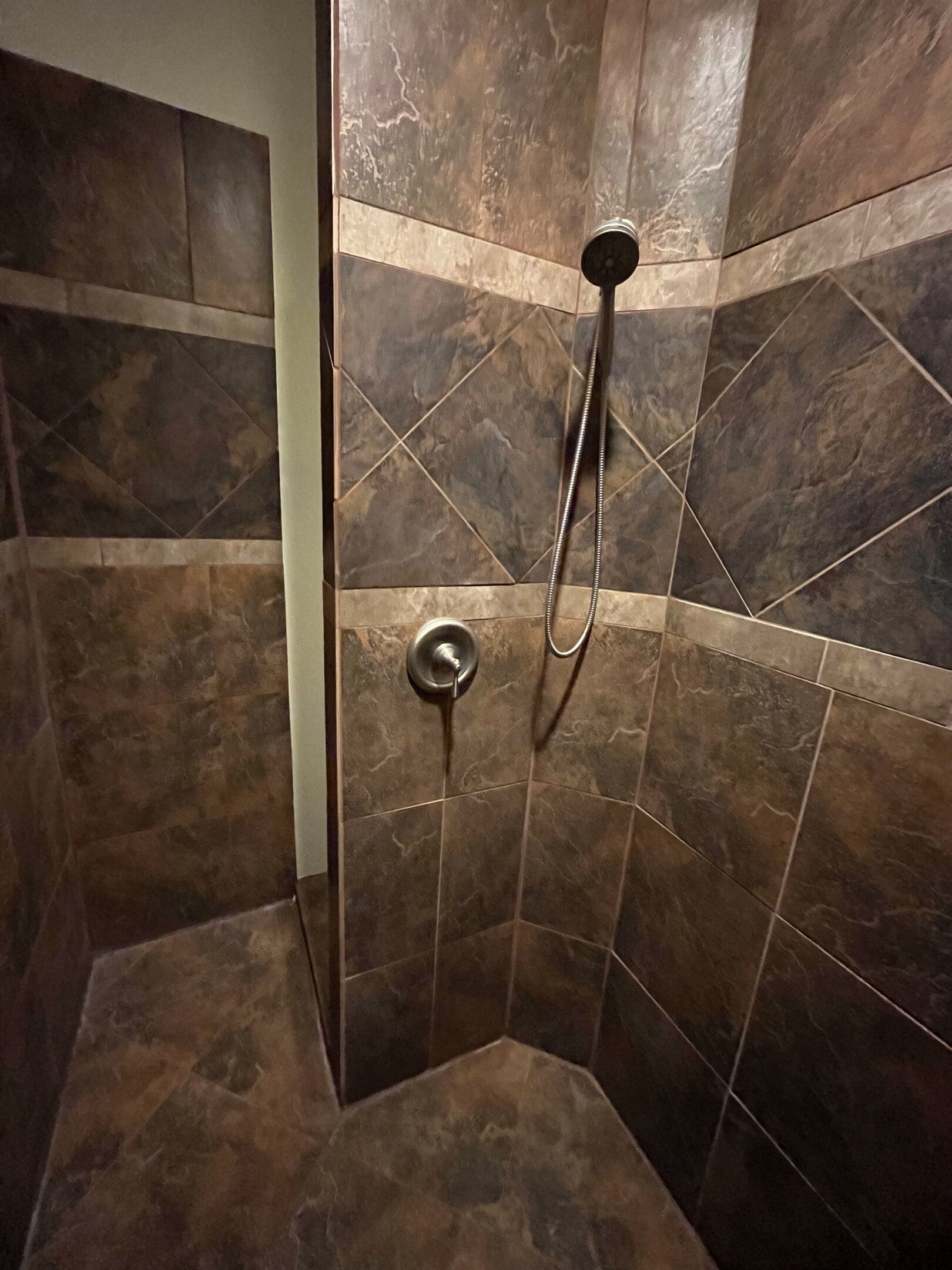 Chuveiro com chuveiro manual e torneira fixada na parede, instalado num canto com azulejos castanhos.