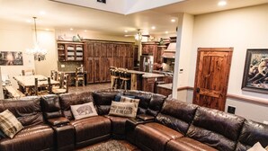 A spacious living room with a large L-shaped leather sectional sofa, a dining area with a wooden table and chairs, and a kitchen with wooden cabinets and a bar area.