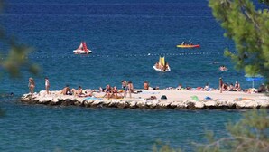 Una playa con gente tomando el sol y nadando, barcos y un muelle con sombrillas.