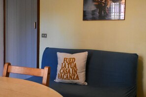 Une pièce avec un canapé bleu, une chaise en bois, un coussin blanc orné d’un motif, et un tableau accroché au mur.