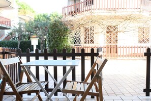 An outdoor seating area with a table and chairs, a wooden fence, and a building with a balcony in the background.