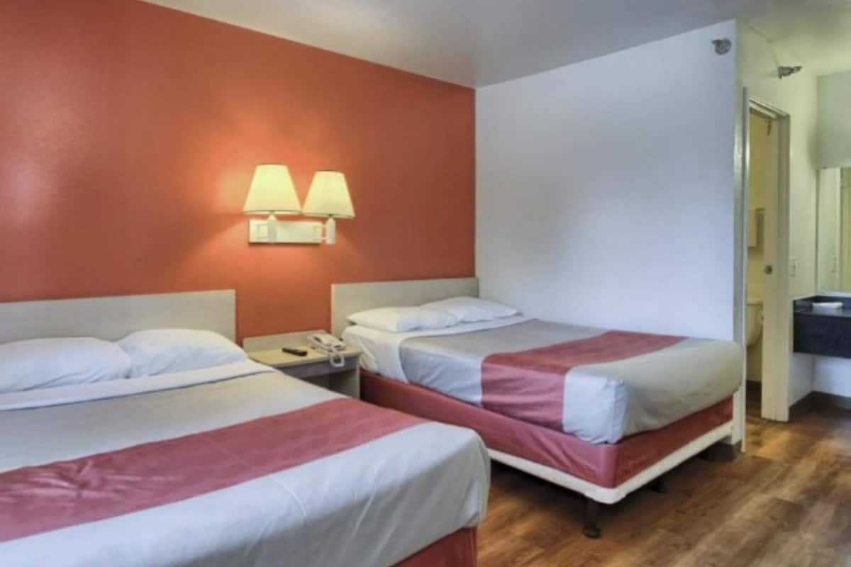 Et hotelværelse med to senge, begge med hvide sengetæpper og røde detaljer, et natbord med en telefon og en lampe monteret på væggen.