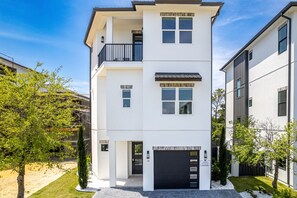 Un immeuble résidentiel moderne à deux étages, doté d’une porte de garage noire, d’un balcon à l’étage supérieur et d’une pelouse bien entretenue agrémentée de quelques arbres.