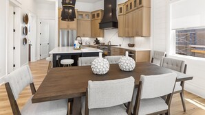 A modern kitchen with wooden cabinets, a central island, and a dining area with a dark wood table and grey upholstered chairs. - Easy Street | New Home | 7 Beds | Pool | Golf Cart | Bikes (Santa Rosa Beach)