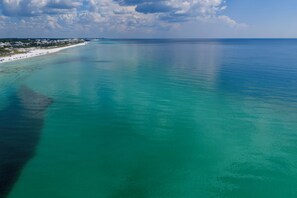 An aerial view of a coastal area with clear turquoise waters, a sandy beach, and a distant shoreline with buildings.
