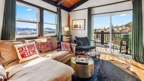 Ein gemütliches Wohnzimmer mit einer Couch, einem Sessel und einem Couchtisch. Große Fenster bieten einen Blick auf die Berge und einen Balkon.