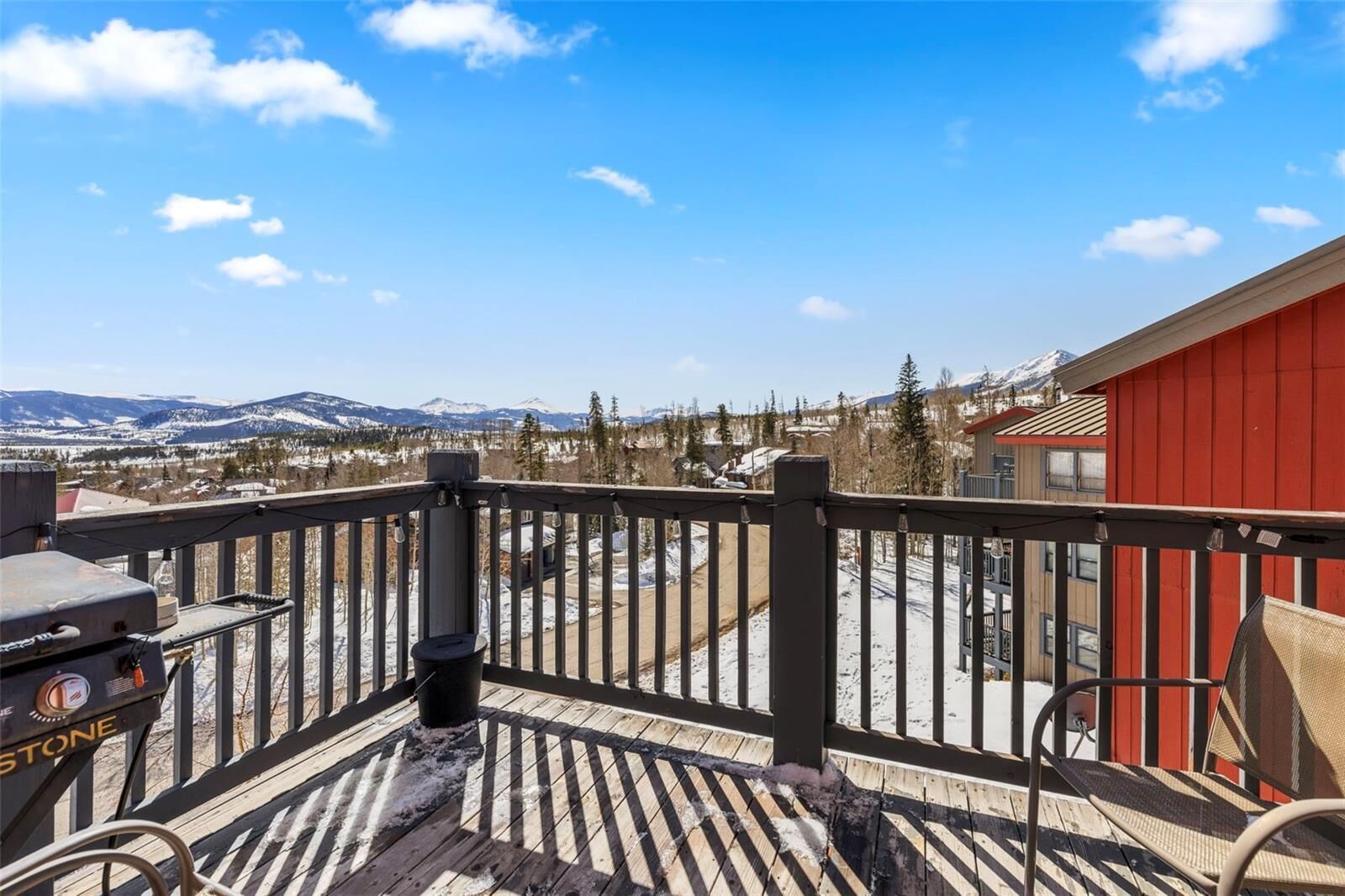 Eine Holzdachterrasse mit Grill und Stühlen, mit Blick auf eine verschneite Landschaft mit Bergen und einem roten Gebäude.