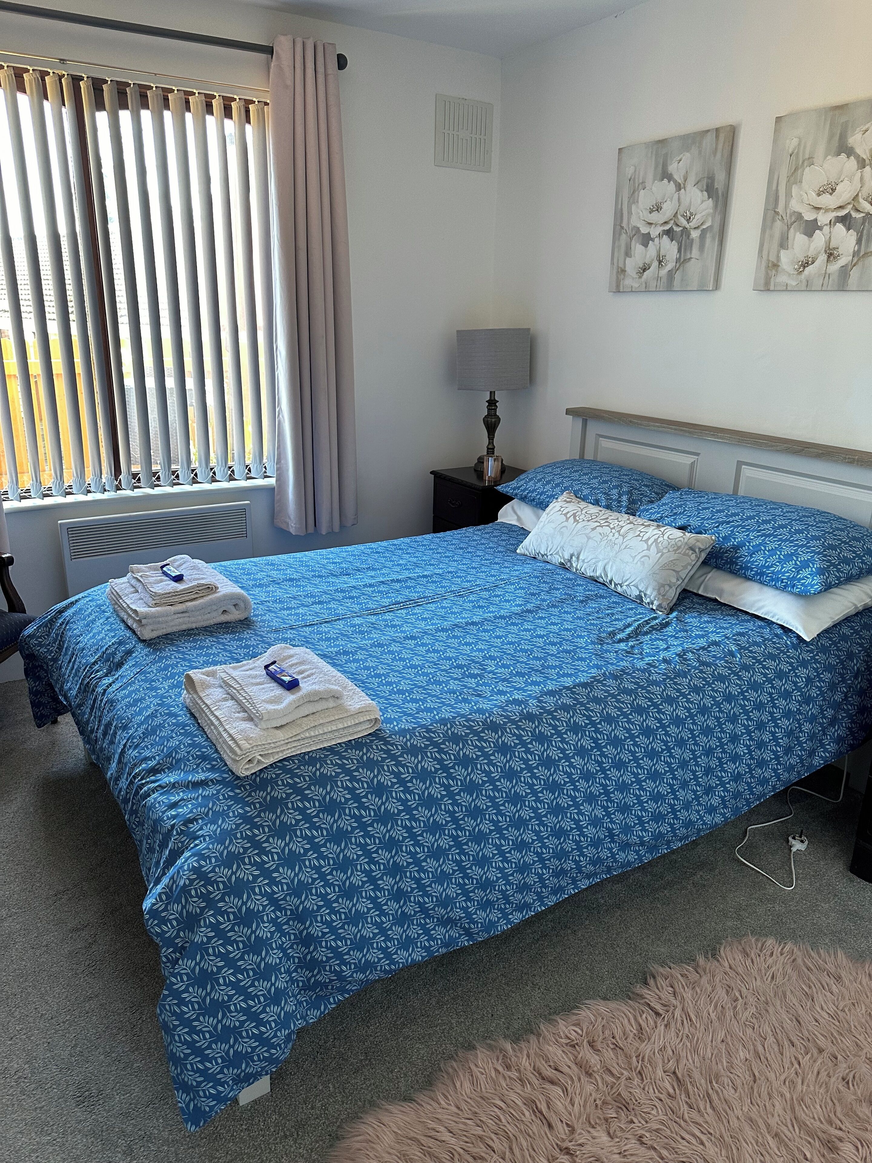 Una cama bien tendida con ropa de cama floral azul, una lámpara de noche y una ventana con persianas.