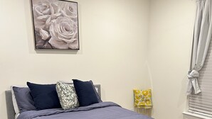A bedroom with a bed, pillows, a framed picture on the wall, a window with curtains, and a small table with a decorative item.