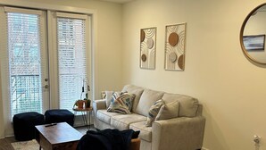 A modern living room with a beige sofa, a wooden coffee table, a black ottoman, and a ceiling fan. There are two abstract wall art pieces and a round mirror on the wall.