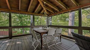 A covered outdoor deck with a wooden table and chairs, surrounded by glass panels and a view of trees. - Toccoa Time - Riverside | Pet Friendly | Firepit | Pool table (Blue Ridge)