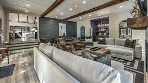 A spacious living room with a large sectional sofa, armchairs, and a coffee table. There is a fireplace, a chandelier, and a decorative deer head on the wall. - Toccoa Time - Riverside | Pet Friendly | Firepit | Pool table (Blue Ridge)