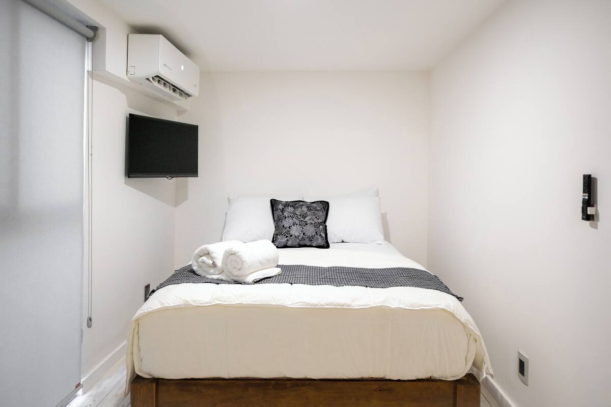 Un lit bien fait, recouvert d’une couverture blanche et grise, avec une serviette blanche enroulée et un oreiller noir et blanc. Un téléviseur à écran plat est fixé au mur, au-dessus du lit.