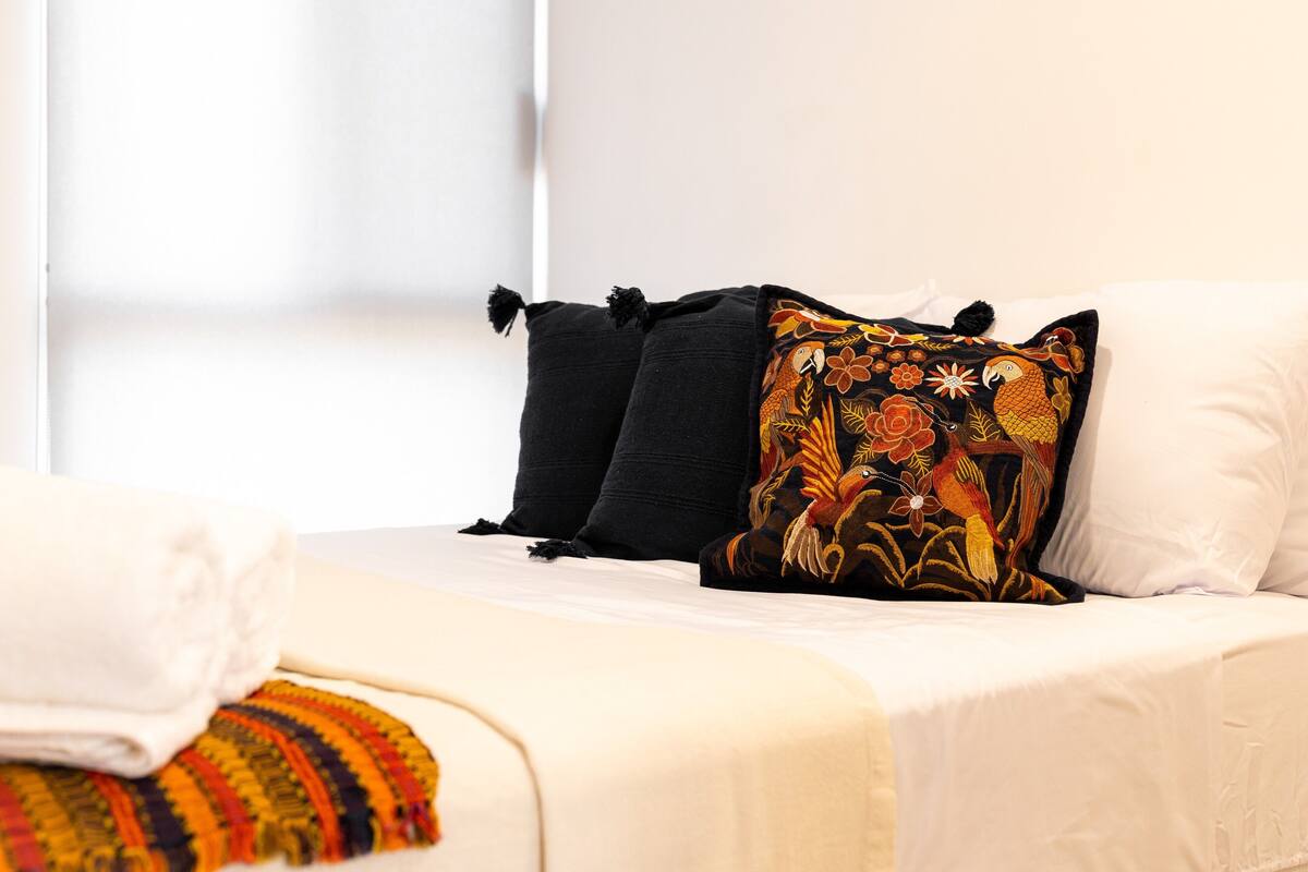 Un lit avec une literie blanche, un coussin brodé aux couleurs vives et un coussin noir.