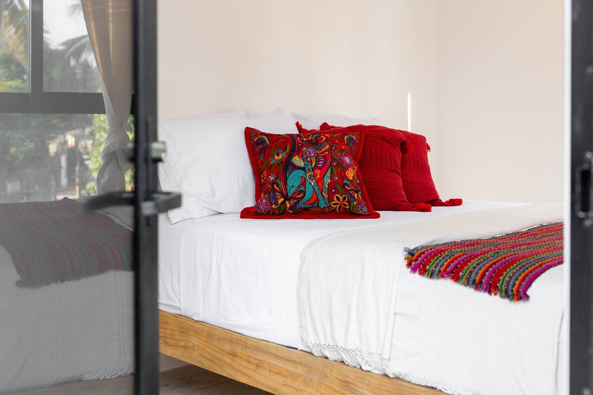 Un lit avec une literie blanche, un coussin coloré brodé et une couverture rouge.