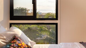 Ein Schlafzimmer mit Bett, Kissen und einem Fenster mit Blick auf Strand und Grünfläche.
