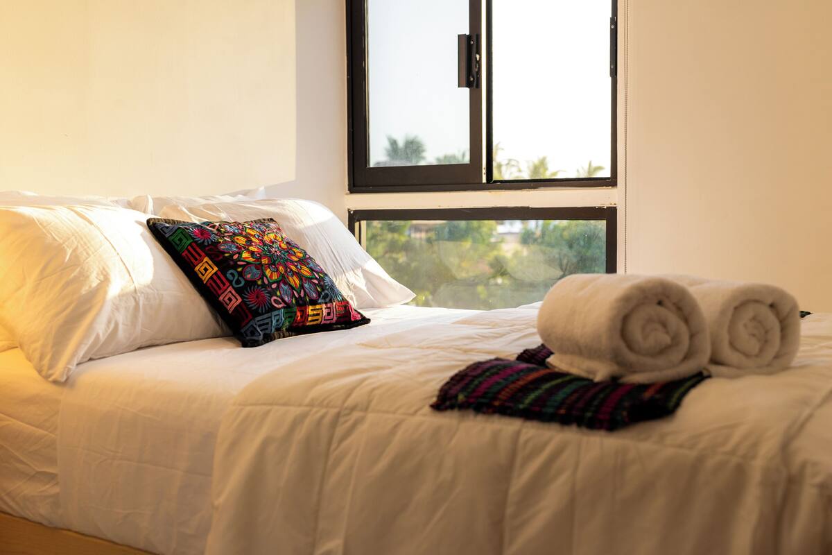 Un lit bien fait, avec des draps blancs, un coussin coloré et une serviette enroulée.