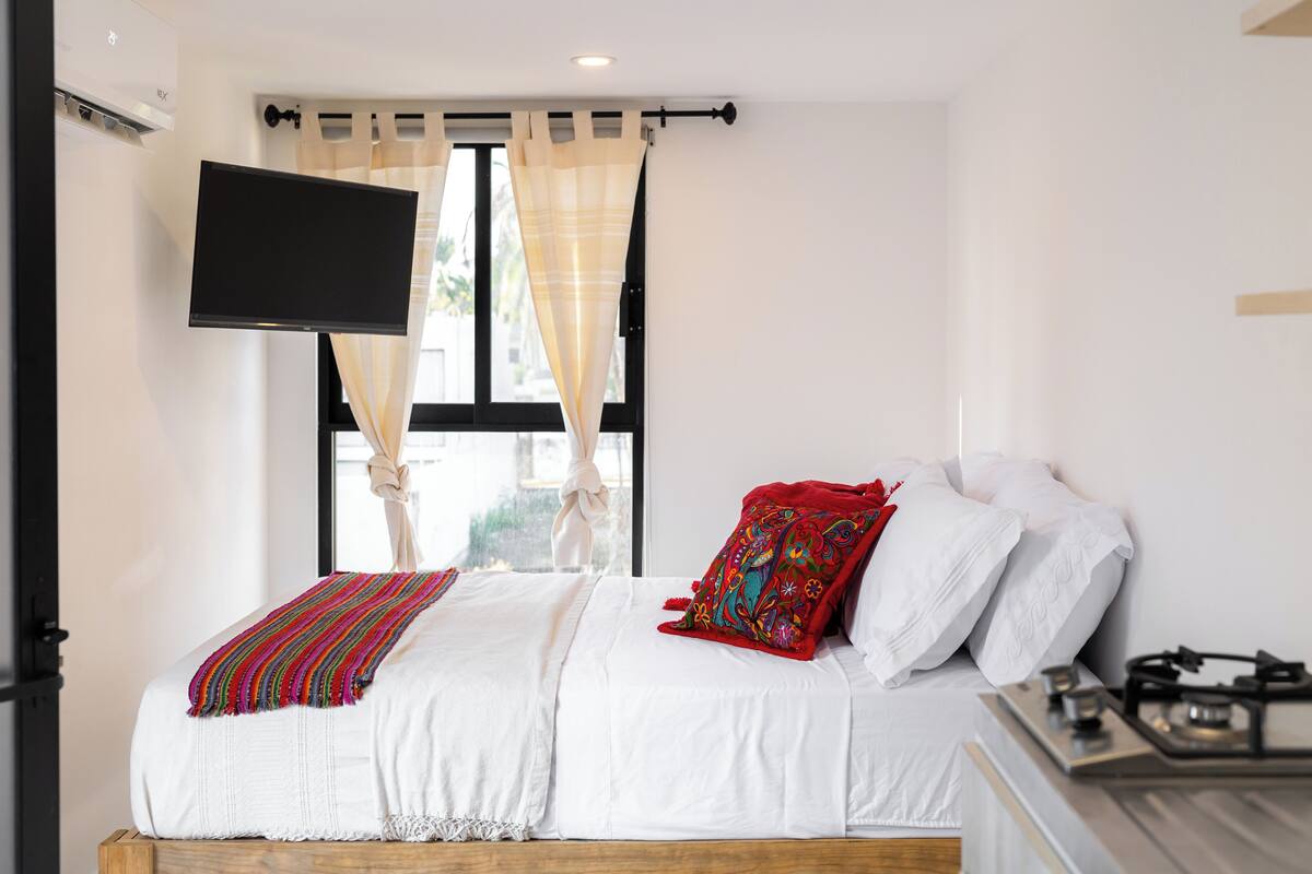 Une chambre à coucher comprenant un lit, un téléviseur à écran plat fixé au mur, une fenêtre avec des rideaux et une cuisinière à gaz.