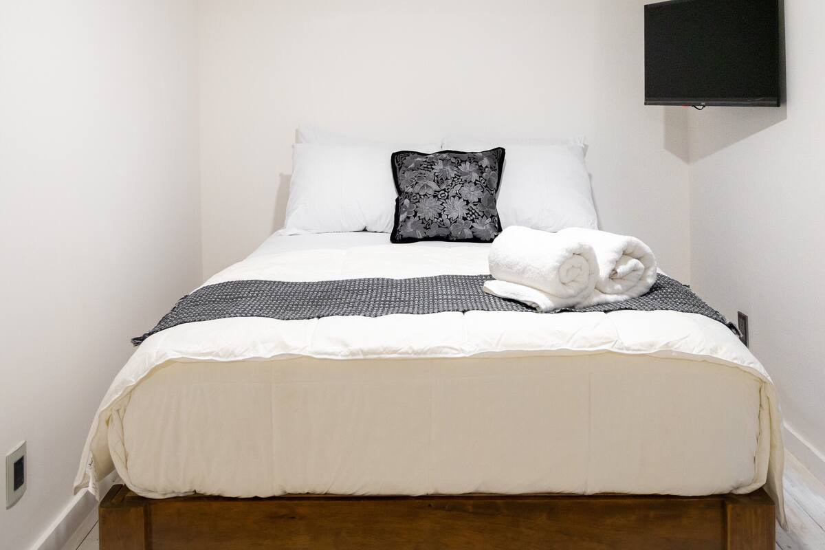 Un lit bien fait, avec une literie blanche et grise, un coussin noir à motifs floraux et une serviette blanche roulée.