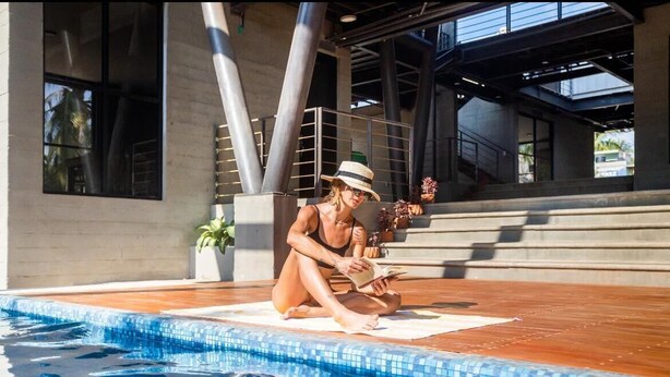 Eine Person sitzt an einem Pool mit Fliesenbord, einer Topfpflanze und einer Treppe im Hintergrund.