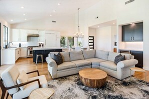 A modern living space with a sectional sofa, a round coffee table, and a kitchen area with white cabinetry. - Golf Paradise: Fairway Views & Private Poolside (La Quinta)