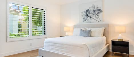 Un dormitorio con una cama grande, dos mesitas de noche, una ventana con persianas y una obra de arte en la pared.