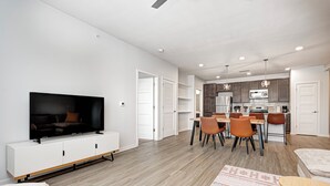 A modern living room with a flat-screen TV, a dining area with a wooden table and chairs, and a kitchen with wooden cabinets and a microwave. - Comfy Cozy - 2King bed/2bth Fishers (Fishers)