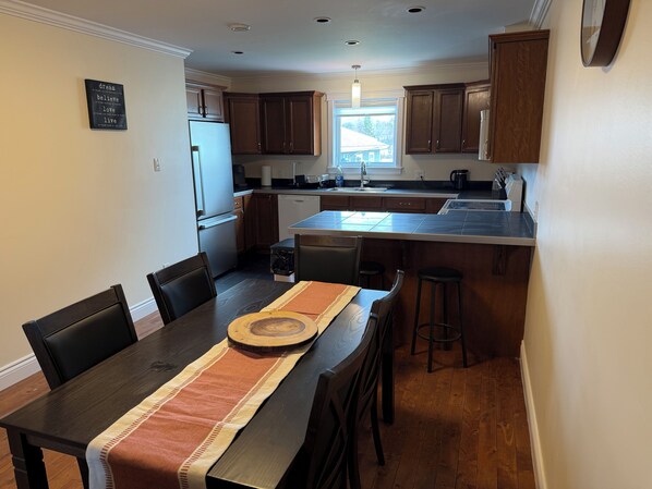 A modern kitchen with dark wooden cabinets, a central island, and a dining area with a table set for four. - The Comfort Loft (Saint John)