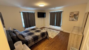 A bedroom with a bed, a TV, a window with curtains, a painting, and a wooden floor.
