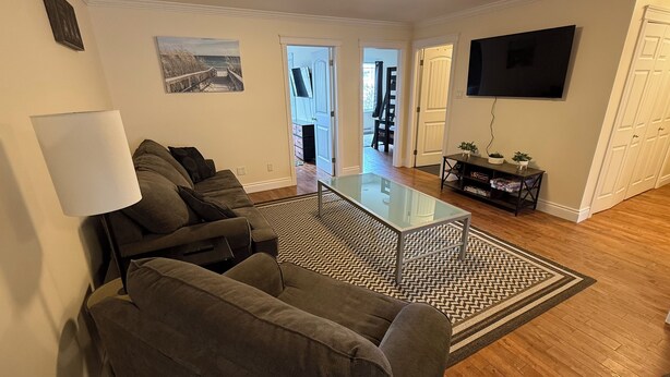 A living room with a sofa set, a glass coffee table, a patterned rug, a TV, and a door leading to another room.