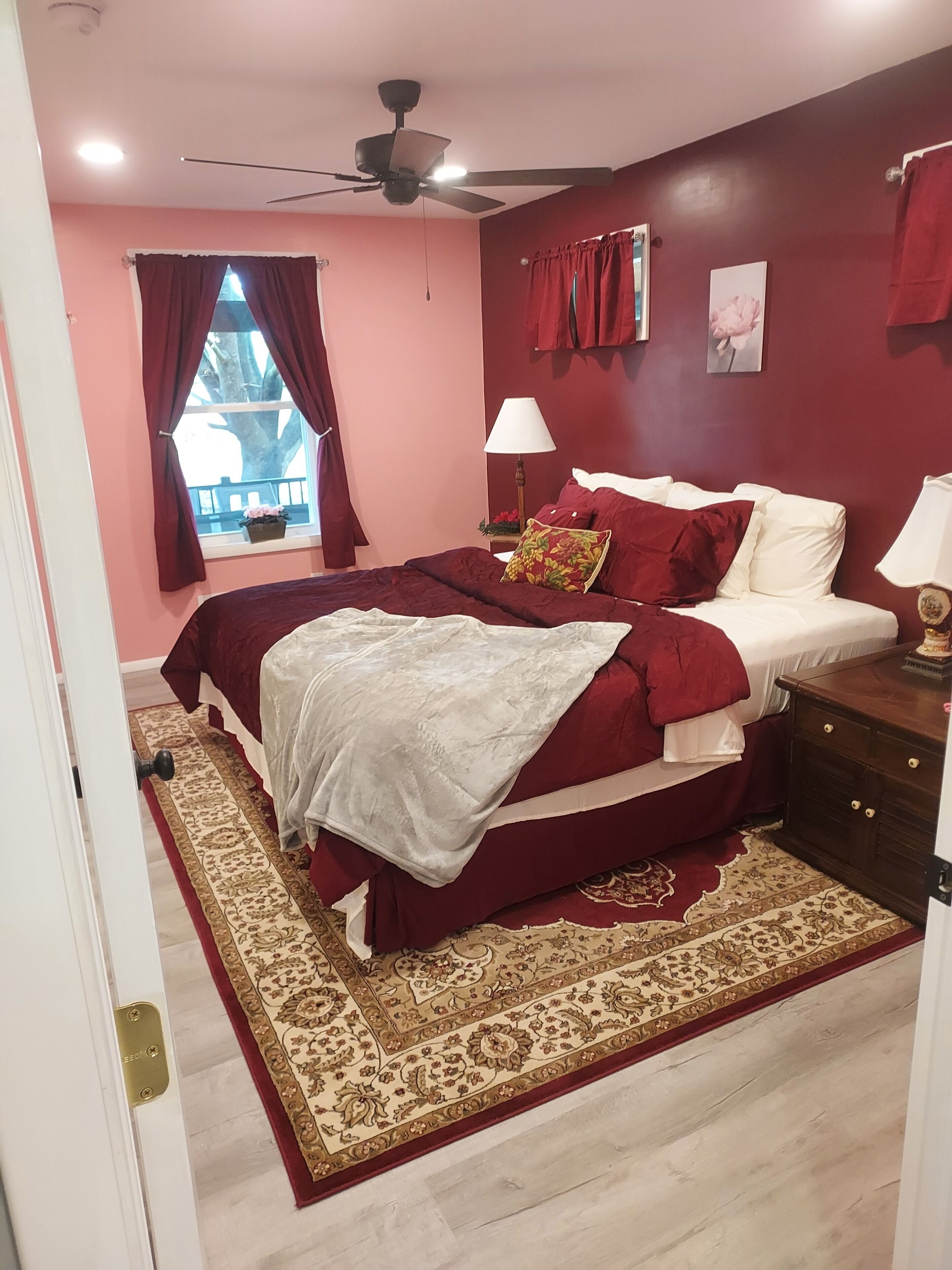 Quarto com cama, mesa de cabeceira, luminária, ventilador de teto, janela com cortinas e um ornamento floral na parede.