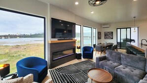 A modern living room with a fireplace, a large TV, and a view of a body of water. - Spectacular Bridge and Bay Views! Bayfront, Hot Tub, Dog Friendly, Fireplace! (Waldport)