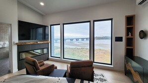 A modern living room with a large window, a flat-screen TV, two brown armchairs, a coffee table, and a fireplace. - Spectacular Bridge and Bay Views! Bayfront, Hot Tub, Dog Friendly, Fireplace! (Waldport)