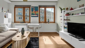 Un salon moderne comprenant un canapé, une table à manger avec des chaises, un téléviseur posé sur un meuble de télévision blanc, et de grandes fenêtres offrant une vue sur la ville.