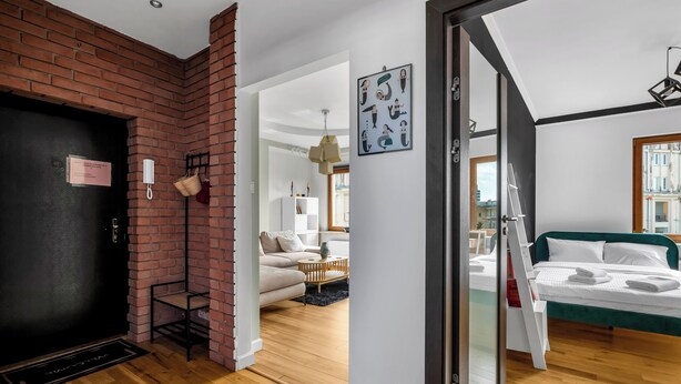Une chambre d’hôtel moderne avec un mur d’accent en briques, un plancher en bois, un lit avec du linge de maison blanc, une tête de lit verte et une vue sur la ville par la fenêtre.