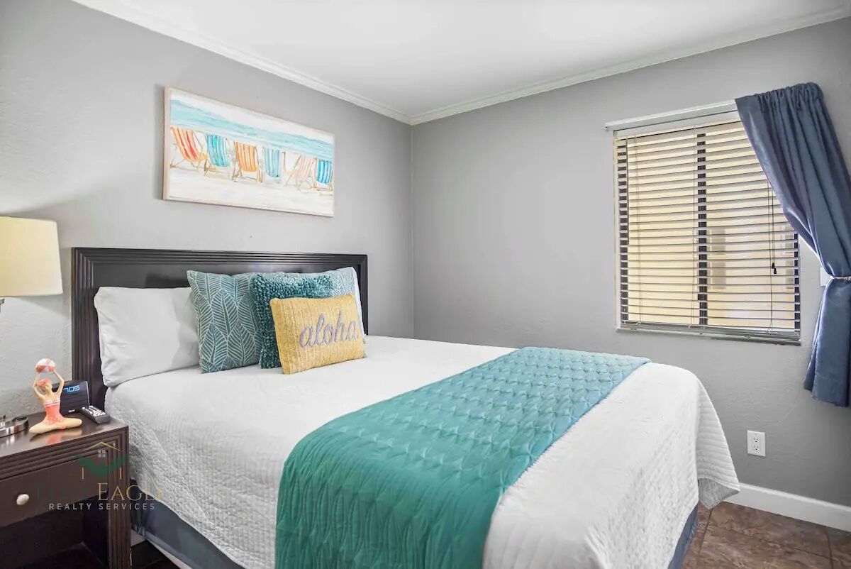 Een slaapkamer met een bed, nachtkastje, een kunstwerk aan de muur en een raam met blinds.