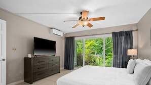 A bedroom with a bed, a TV on a dresser, a ceiling fan, and a view of greenery through the window. - 2br 2ba Modern Kamahana, Pool, Close to Beach (Princeville)