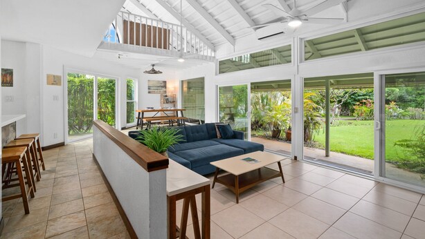 A spacious living area with a blue sofa, a coffee table, and a dining table with chairs. There is a balcony with a railing and a view of the garden. - 2br 2ba Modern Kamahana, Pool, Close to Beach (Princeville)